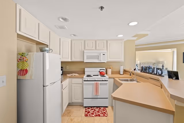 A clean and well-lit kitchen with white cabinets, a white refrigerator, a white stove with a red kettle on it, a microwave above the stove, and beige countertops. There is a red and white patterned rug in front of the stove and a pink towel hanging on the oven handle. The kitchen opens to a living area with a decorative sign and a lamp visible in the background.
