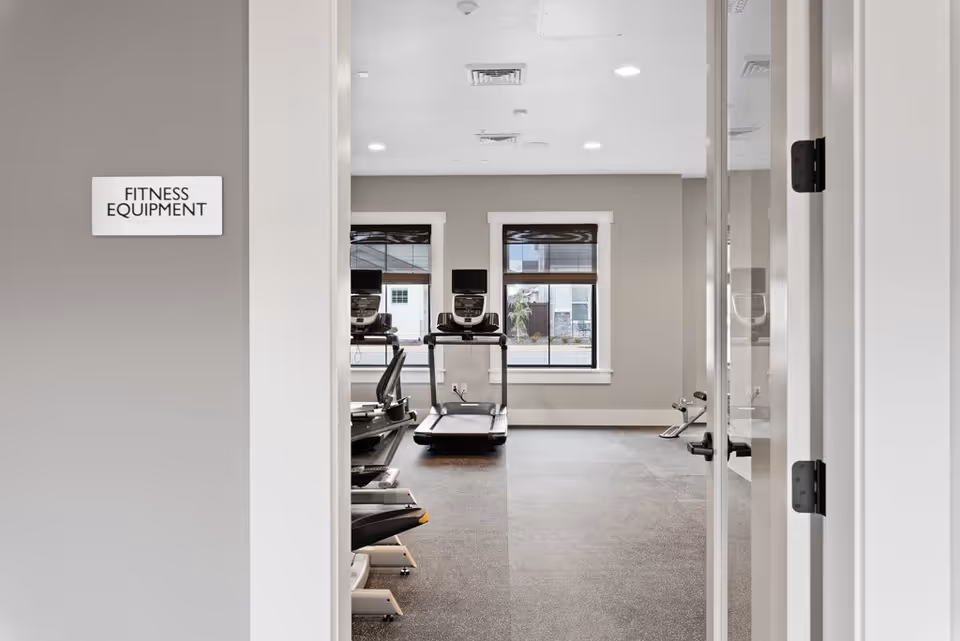 View through an open doorway into a fitness equipment room with treadmills and exercise bikes. The room has two windows with blinds, gray walls, and a speckled rubber floor. A sign on the left wall outside the room reads 'FITNESS EQUIPMENT'.