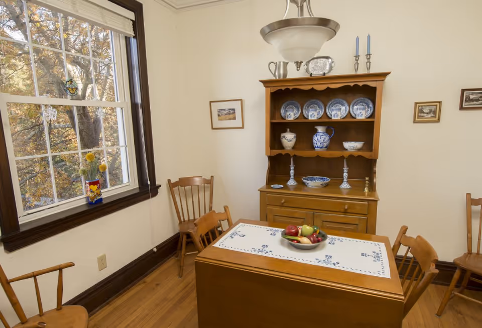 A cozy dining room with a wooden table covered by a white and blue patterned table runner. On the table is a bowl filled with various fruits. Surrounding the table are wooden chairs. Against the wall is a wooden hutch displaying blue and white china plates, pitchers, and bowls. A window with white blinds lets in natural light, showing trees outside. The walls are light-colored with small framed pictures hanging.