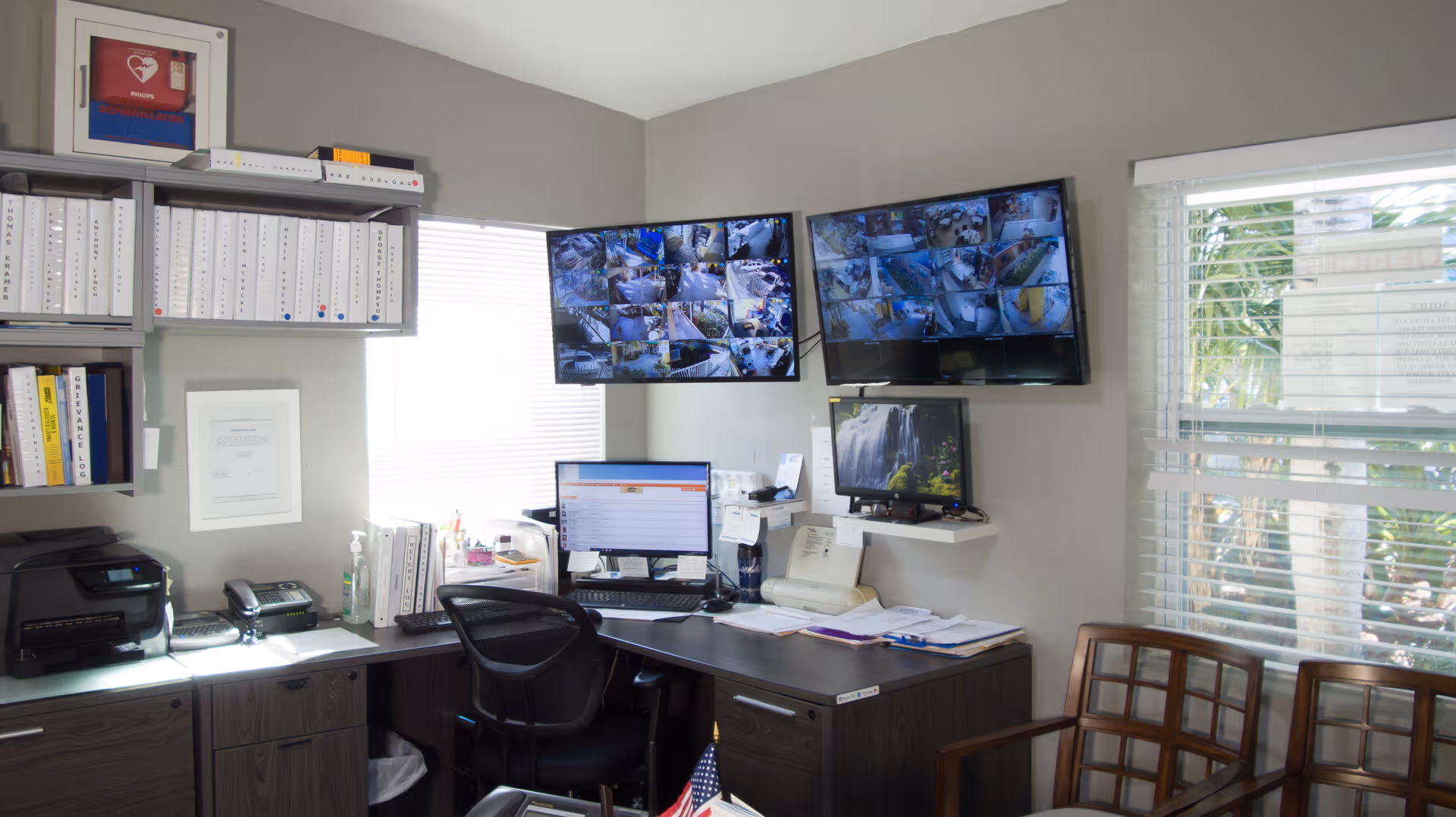 Office room with a desk, computer monitors, and multiple surveillance screens mounted on the wall. The desk has office supplies, a printer, and paperwork. There are windows with blinds allowing natural light into the room.