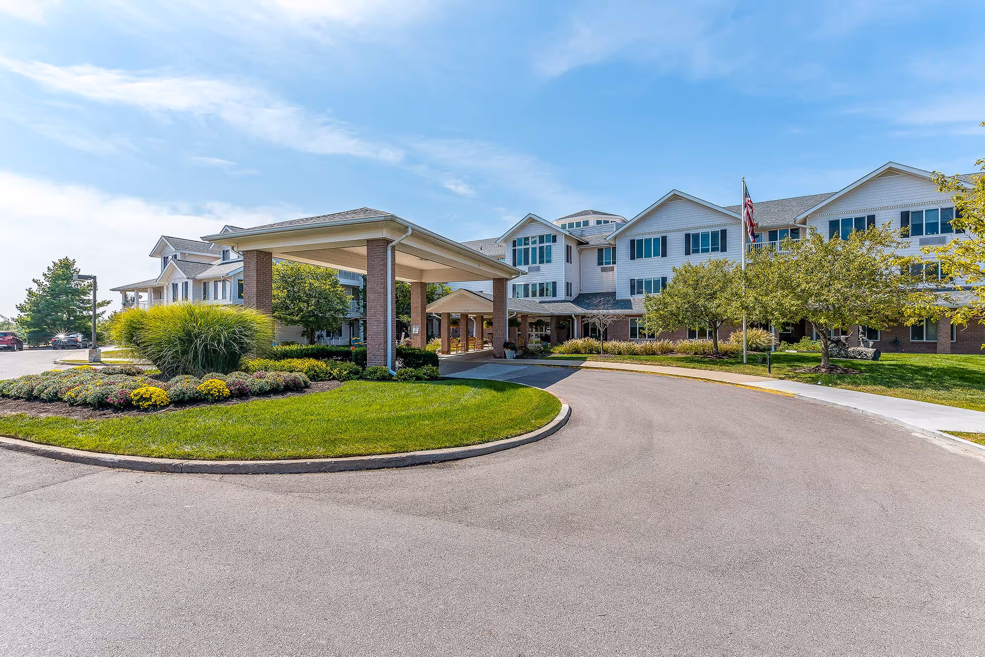 Front entrance of a multi-story senior living building with a covered porte-cochere, circular driveway and landscaped lawn under a blue sky.