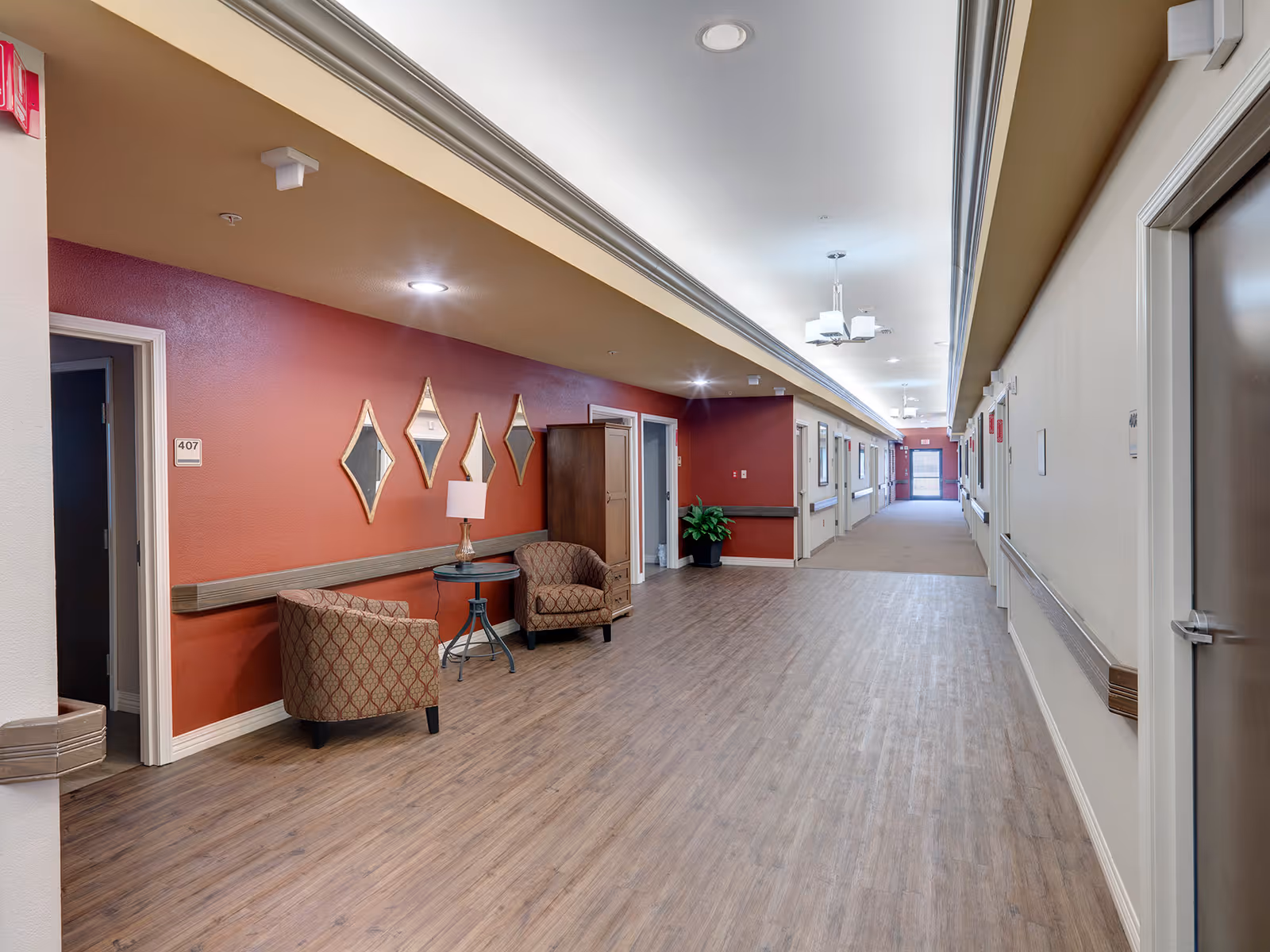 A long, well-lit hallway in a senior living facility with wood flooring and beige and red walls. There are two patterned armchairs and a small round table with a lamp against the red wall, along with four diamond-shaped mirrors. Several doors line the hallway, and a potted plant is visible near the far end.