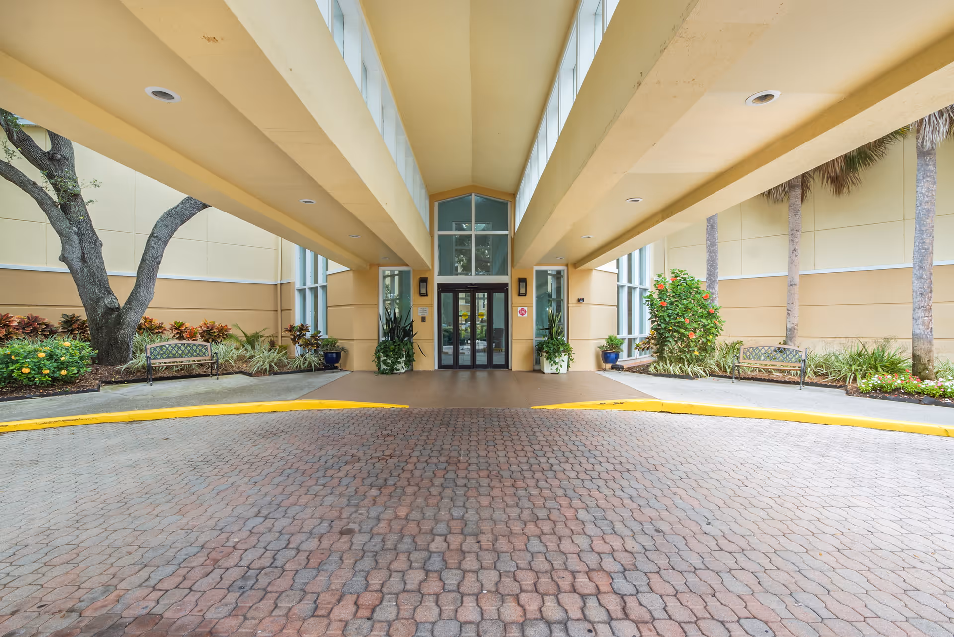 Entrance area of The Barrington facility with a covered driveway, brick-paved ground, glass double doors, potted plants, benches, and landscaping including trees and shrubs.