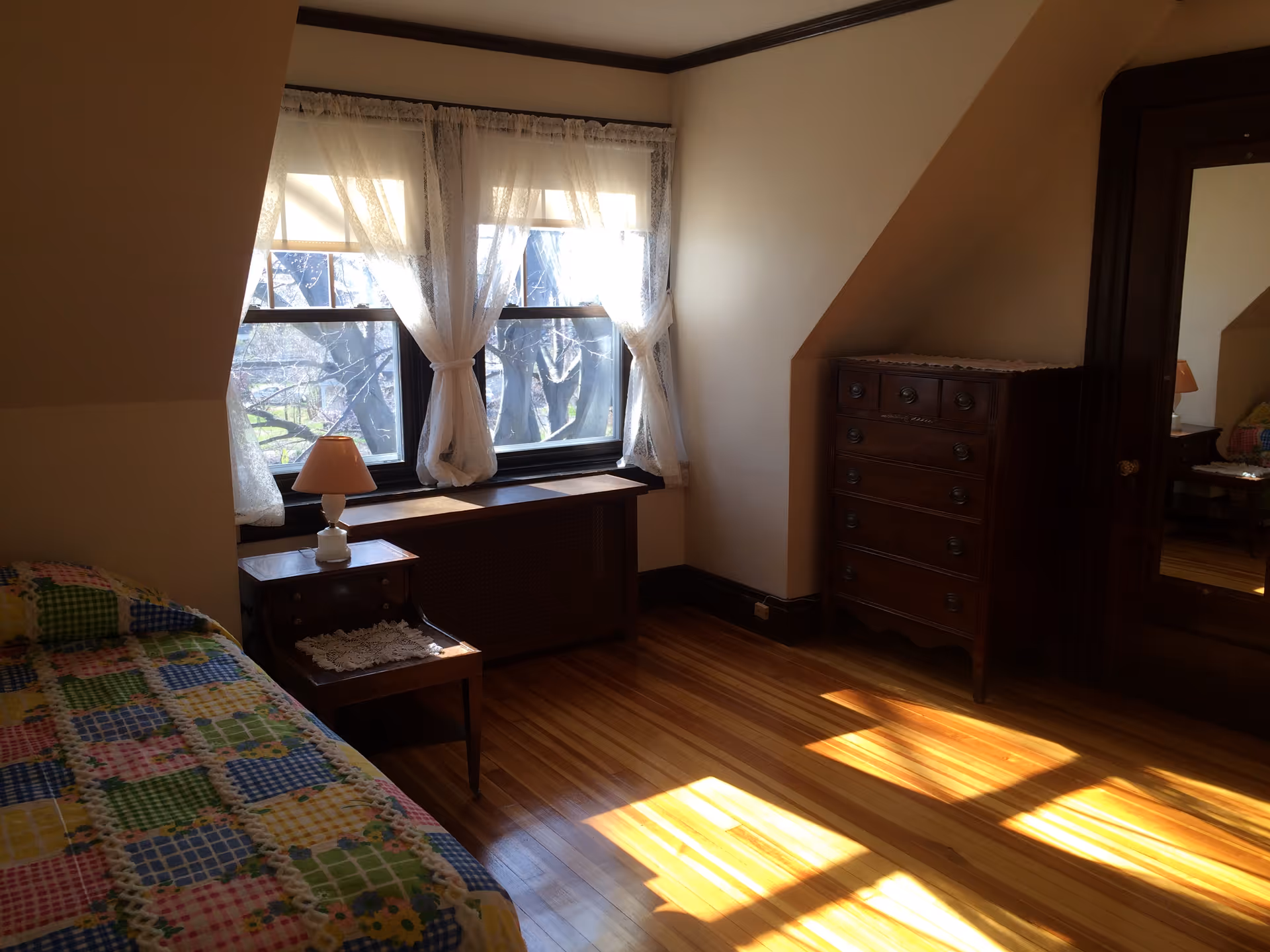 Sunlit bedroom with a bed, nightstand and lamp by a curtained window, wooden dresser and hardwood floors.
