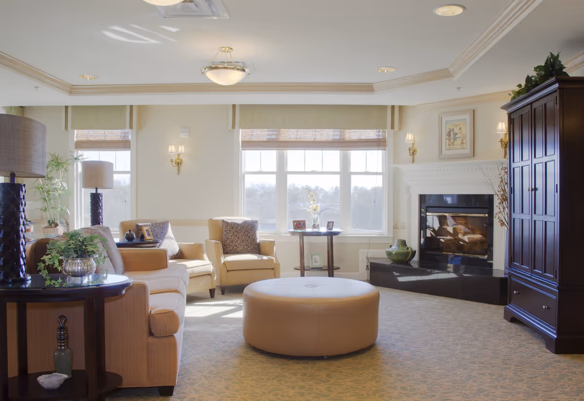 A bright and cozy living room with beige armchairs and a sofa arranged around a large round ottoman. The room features large windows with light green valances, a fireplace with a white mantel, a dark wooden cabinet, and decorative plants and lamps.