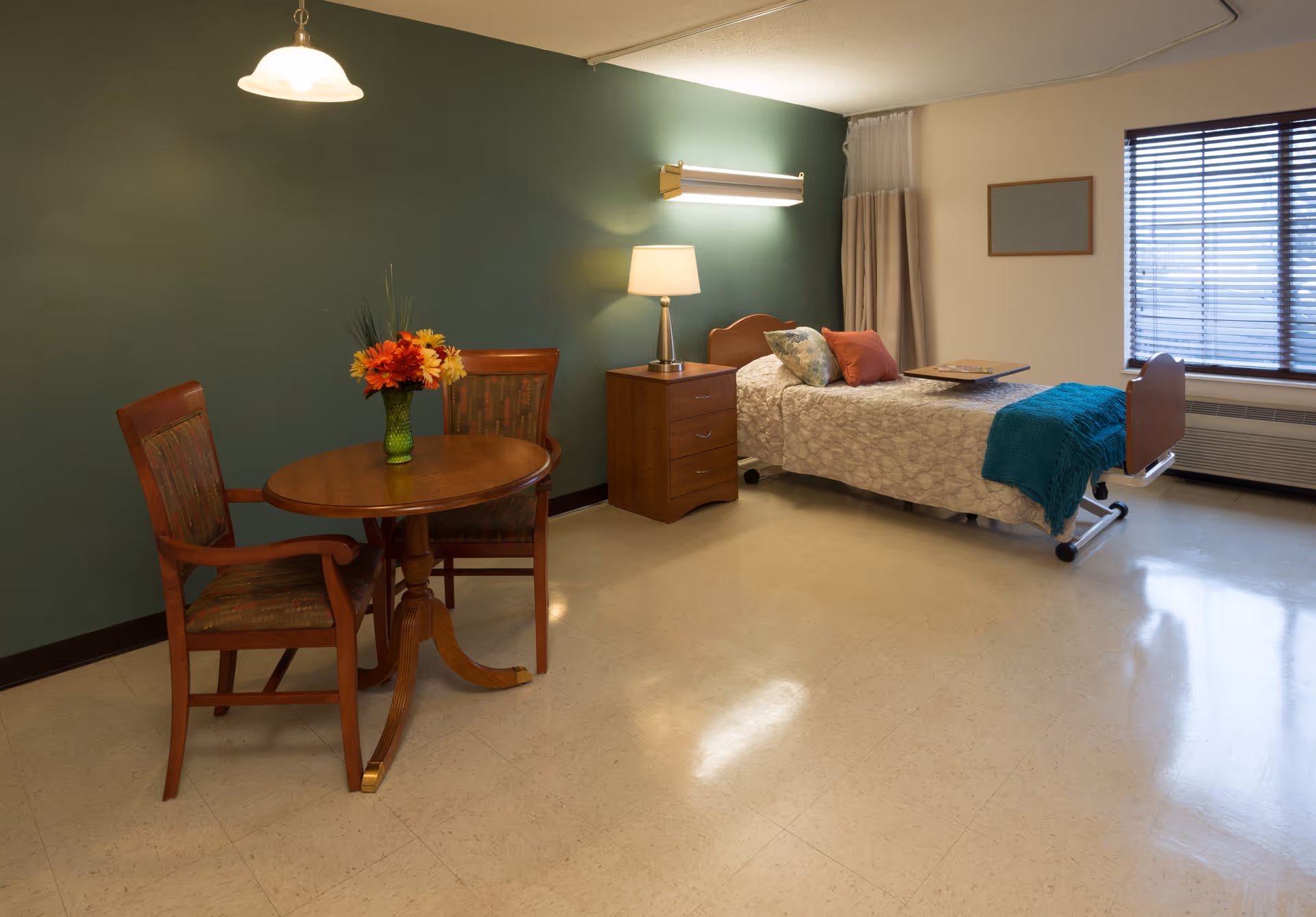 A senior living facility room with a single bed covered in a light patterned bedspread, a teal throw blanket, and decorative pillows. Next to the bed is a wooden nightstand with a lamp. A small round wooden table with two chairs and a vase of flowers is positioned near a green accent wall. A window with blinds and curtains is on the right side of the room.