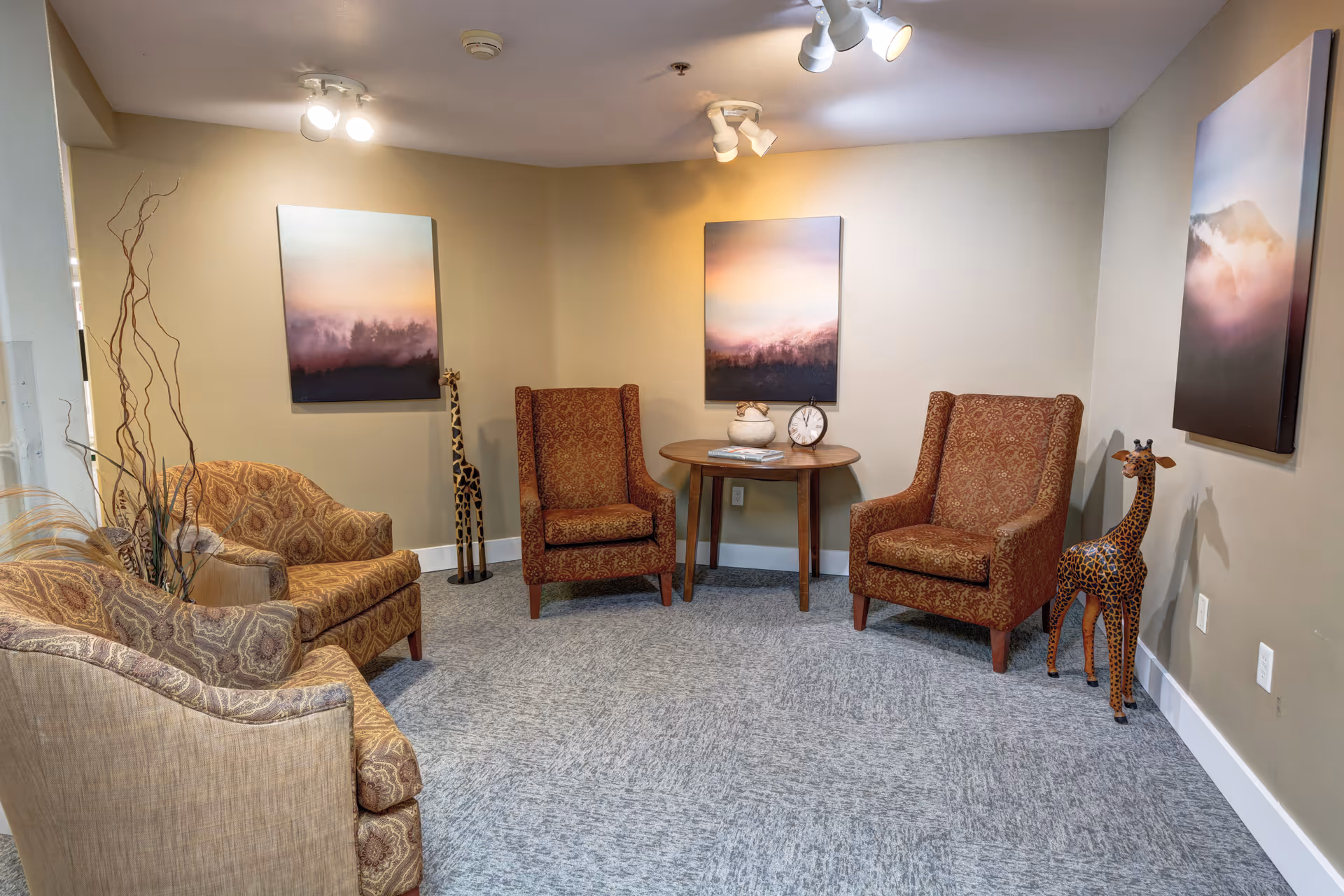 A cozy sitting area with four patterned armchairs arranged around a small wooden table. The table holds a decorative pot, a clock, and some magazines. The walls are beige and decorated with three landscape paintings featuring misty mountains. Two tall giraffe sculptures stand near the corners of the room. The floor is carpeted in a light gray pattern, and ceiling spotlights illuminate the space.