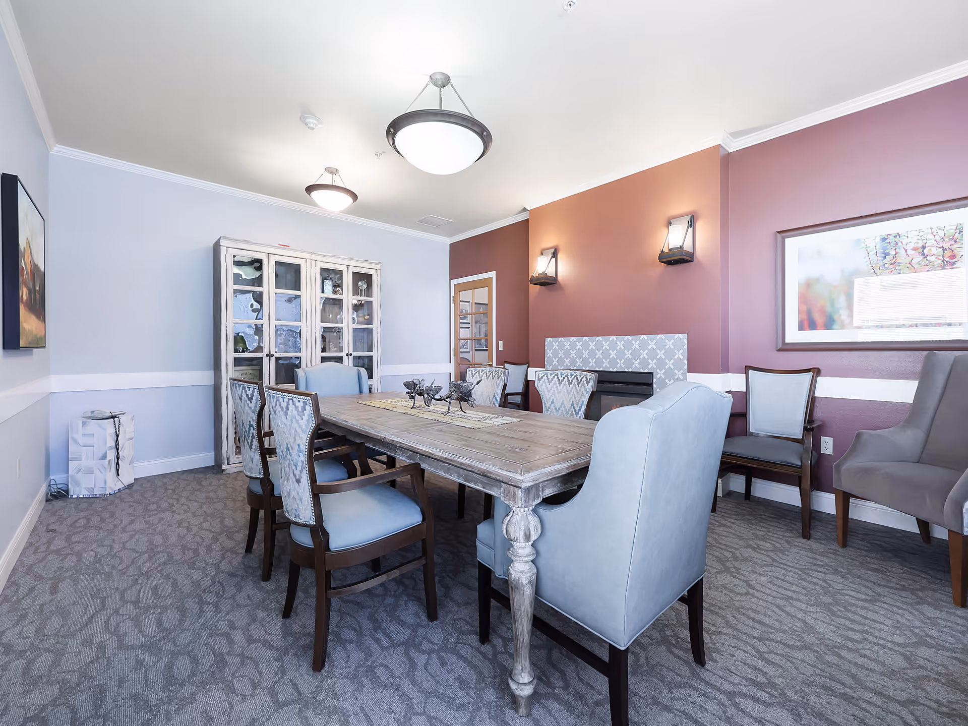 A dining room with a wooden table surrounded by eight upholstered chairs in light blue and patterned fabric. The room has a carpeted floor, a white and purple wall with two wall sconces, a framed picture, and a glass-front cabinet filled with decorative items. Two ceiling lights illuminate the space.