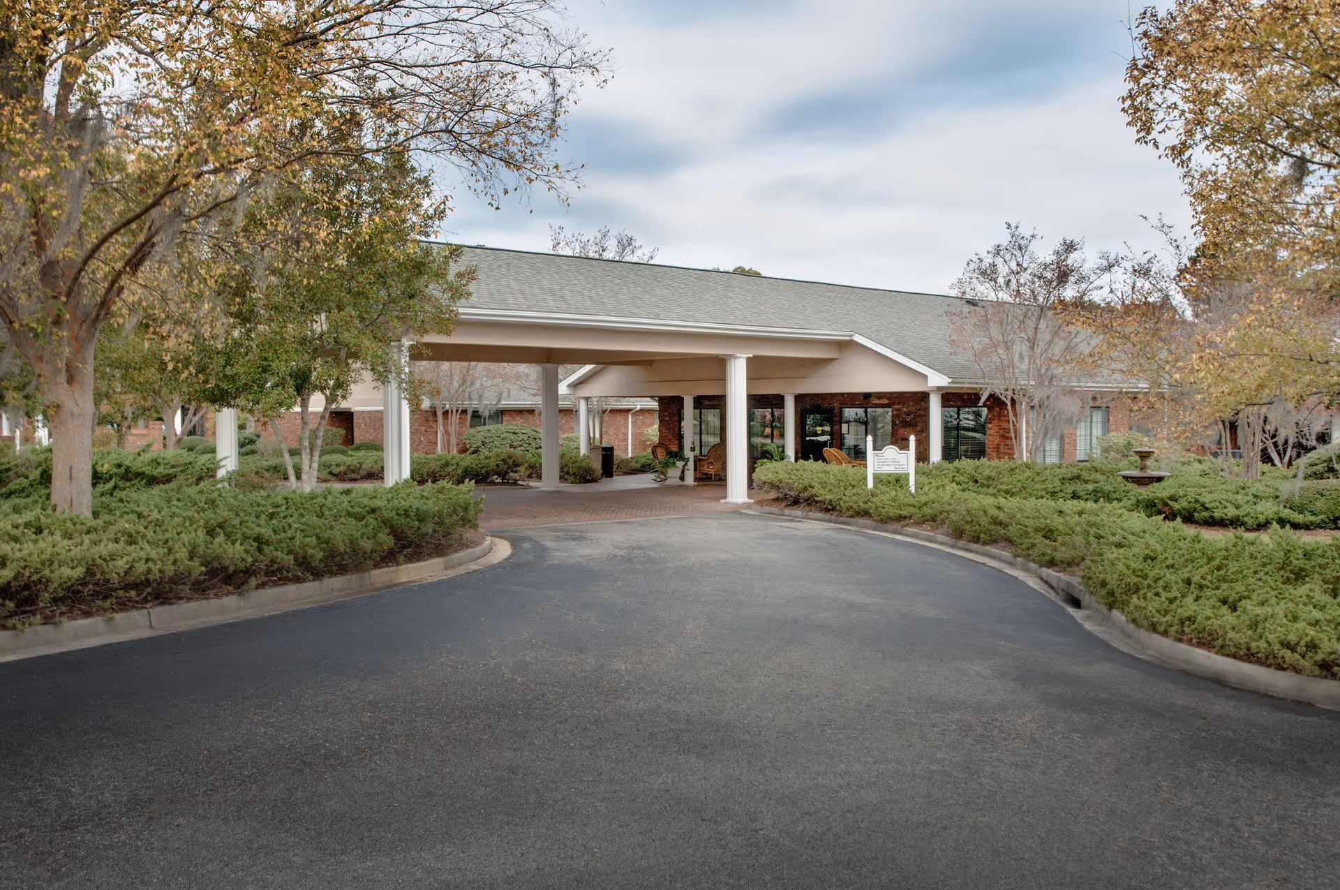 Covered entrance and driveway leading to the front of a single-story brick senior living building surrounded by landscaping and trees.