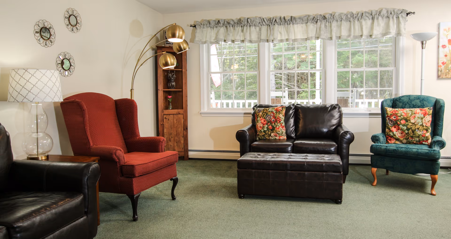 A cozy living room with a green carpet, featuring a black leather loveseat with floral cushions, a red upholstered armchair, a green upholstered armchair with a floral cushion, a brown leather ottoman, a wooden corner shelf with decorative items, a floor lamp with three golden lampshades, a table lamp with a patterned shade, and three decorative round mirrors on the wall. Large windows with white curtains allow natural light to brighten the room.