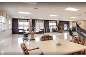 A spacious and well-lit rehabilitation room with large windows, exercise equipment including stationary bikes and parallel bars, a table with floral upholstered chairs, and a tissue box on the table. The room has a clean, polished floor and neutral-colored walls.