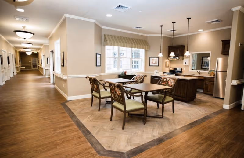 A communal dining nook with a wooden table and four upholstered chairs beside an open kitchen and a long hallway.