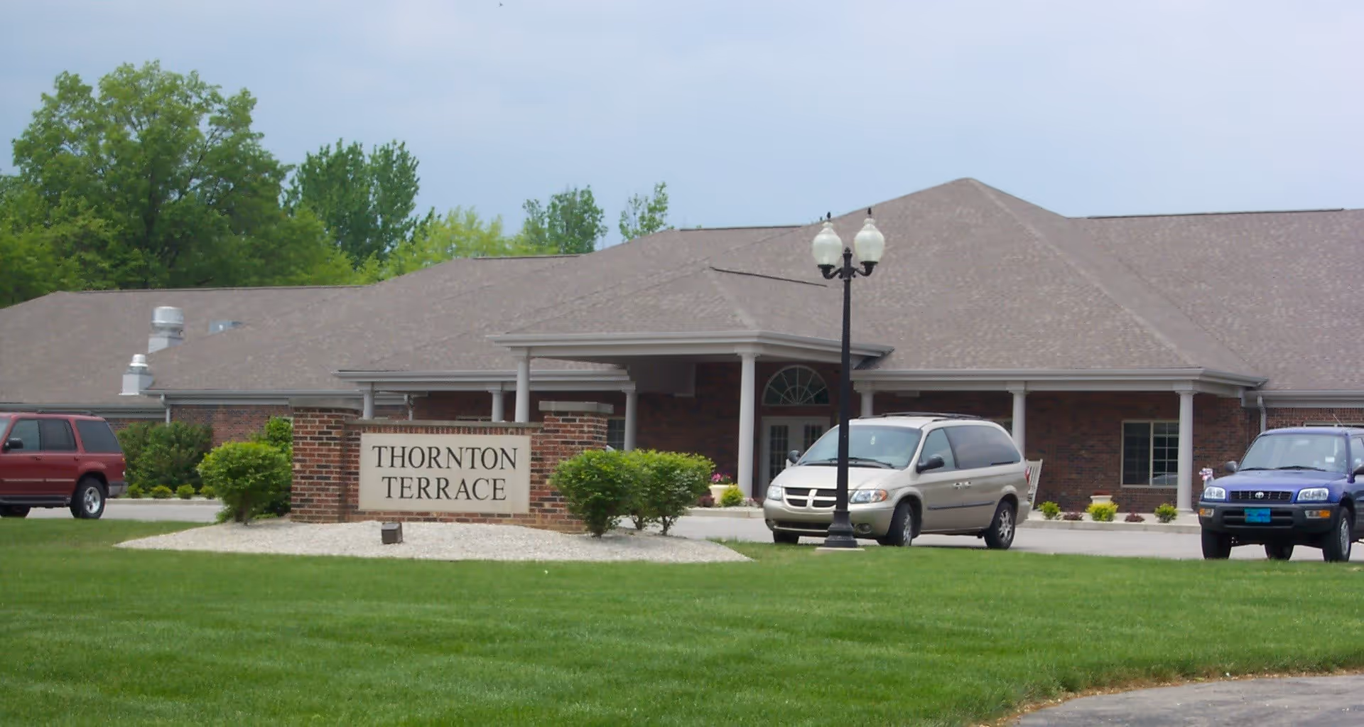 Exterior view of Thornton Terrace Health Campus building with a brick sign in front displaying the facility name. There are three vehicles parked near the entrance, a lamppost, and green grass with trees in the background under a cloudy sky.
