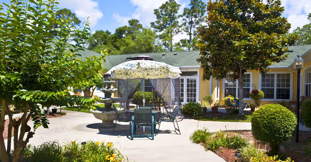 Outdoor courtyard area with a round table and chairs under a floral umbrella, surrounded by trees, bushes, and flowering plants. A multi-tiered water fountain is visible near the table. The courtyard is enclosed by a yellow building with multiple windows and a green roof under a partly cloudy sky.