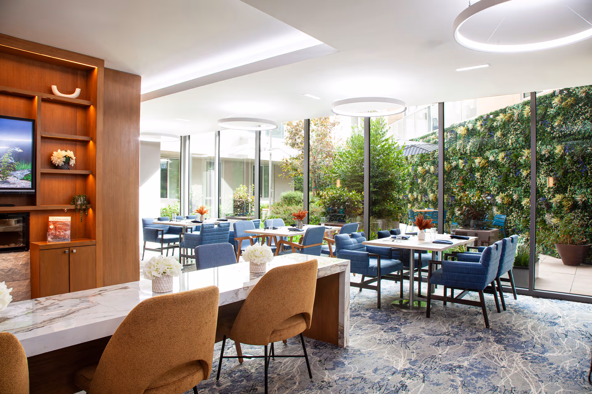 Bright and modern dining area with blue upholstered chairs and wooden tables, large floor-to-ceiling windows overlooking a green vertical garden wall, a marble countertop with beige chairs in the foreground, and circular ceiling lights.