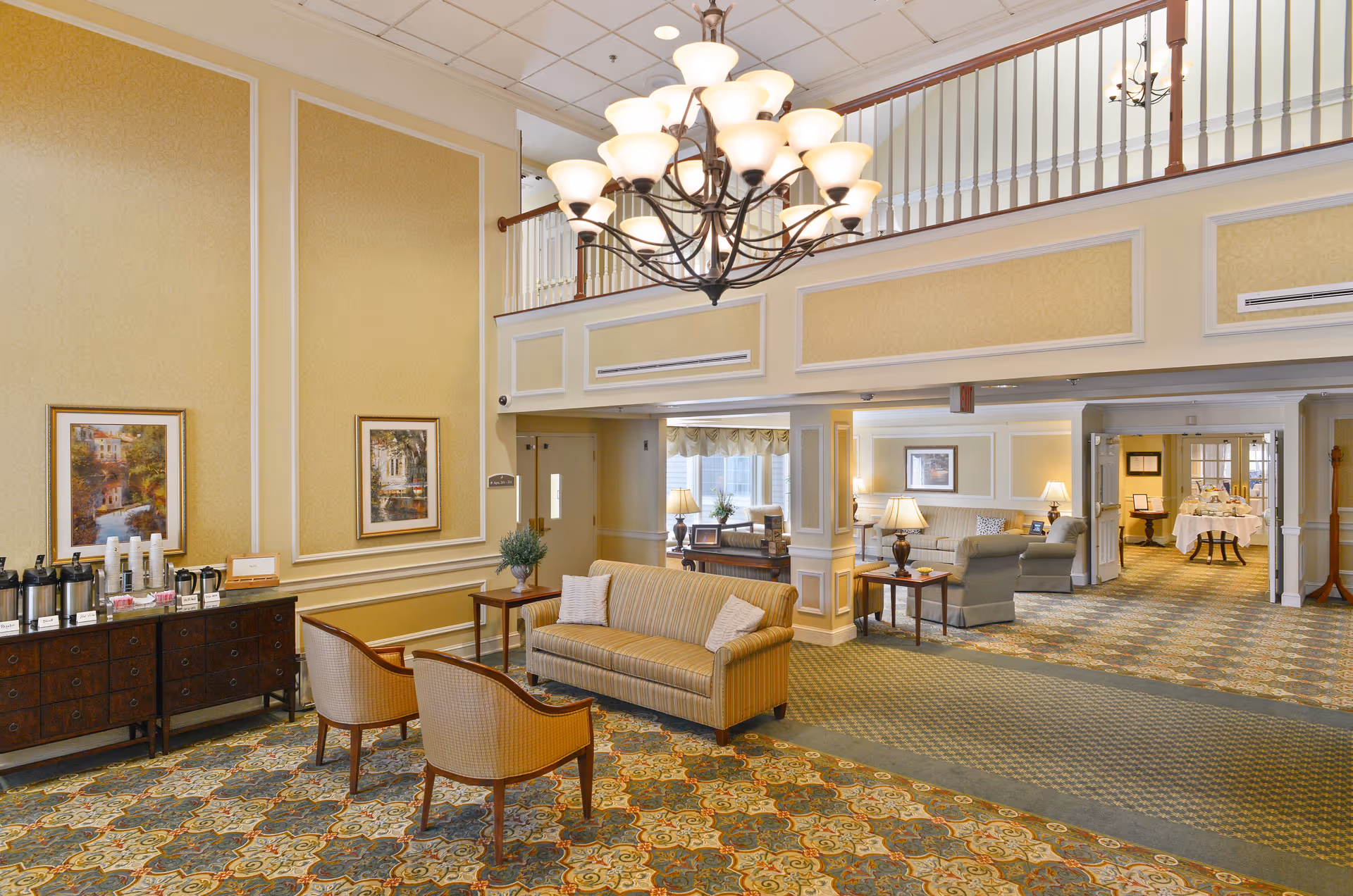 A spacious, well-lit senior living facility lounge with sofas and chairs, a large chandelier, and a coffee station.