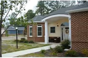 Exterior view of a brick building with a covered entrance supported by white columns, a walkway leading to the door, and some landscaping with small plants and a tree nearby.