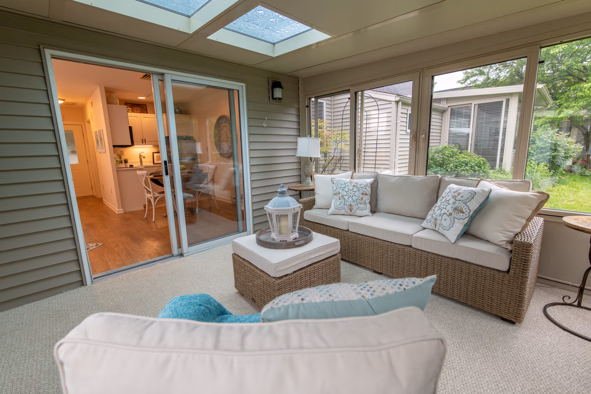 A bright sunroom with large windows showing a garden outside. The room features a wicker sofa with light cushions and decorative pillows, a matching ottoman with a lantern on top, and a cushioned armchair in the foreground. Sliding glass doors lead to an interior space with a kitchen and dining area visible.