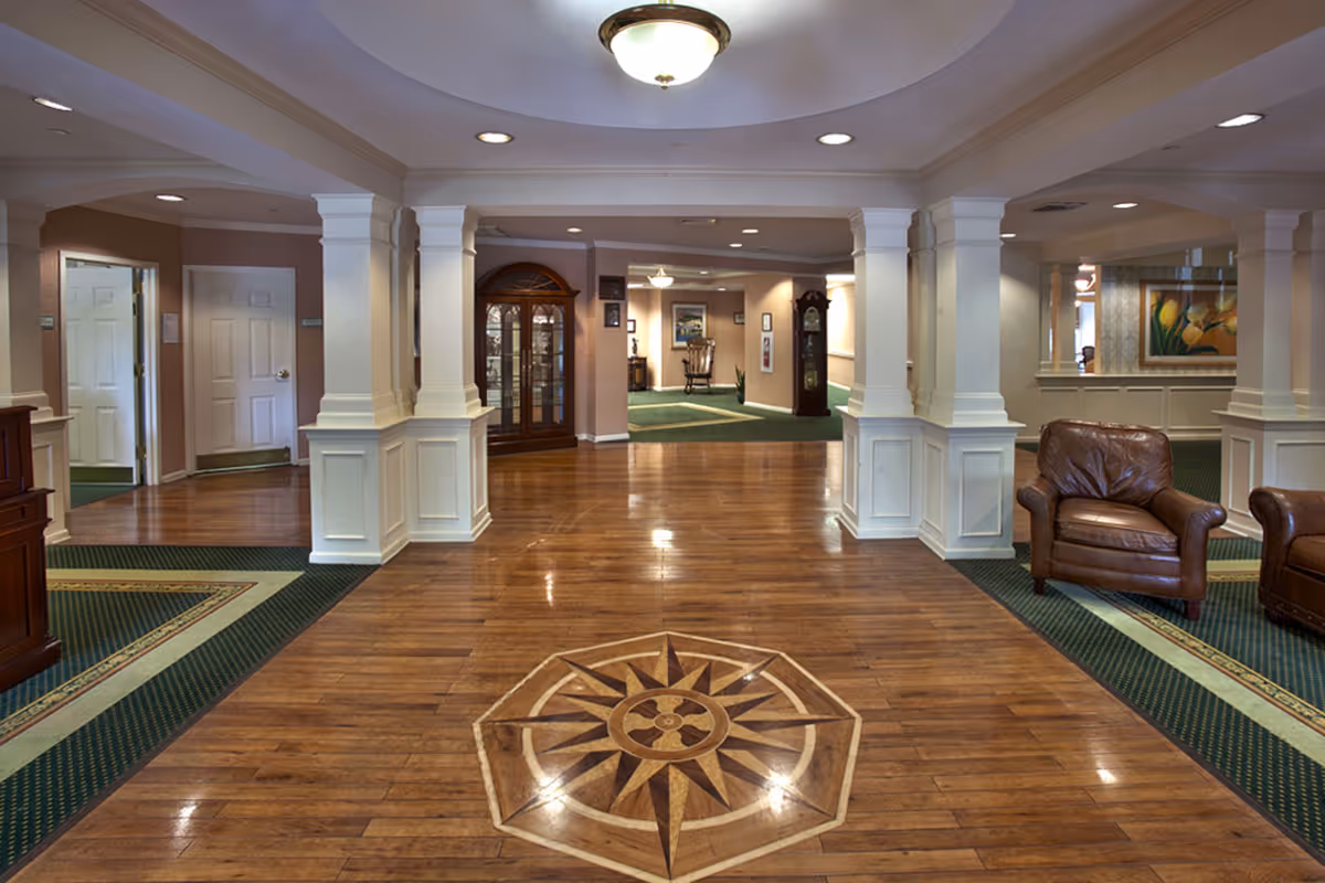 Spacious interior common area with polished wooden floor featuring a decorative compass design in the center. The room has white columns with paneling, recessed lighting, and a ceiling light fixture. There are leather armchairs on the right side, green carpeted areas with patterned borders, a wooden display cabinet, a grandfather clock, and framed artwork on the walls.