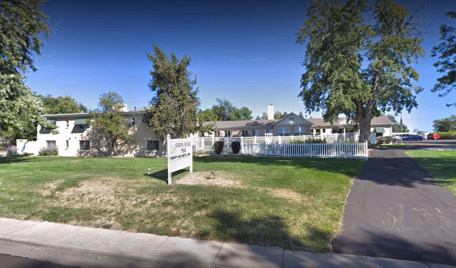 Front exterior of an assisted living building with a lawn, white picket fence, driveway, and a sign.