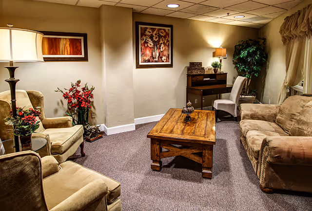 A cozy senior living facility lounge area with beige upholstered armchairs and a sofa arranged around a wooden coffee table. The room features soft lighting from a floor lamp and a table lamp, decorative plants, framed artwork on the walls, and a small desk with a chair in the corner.