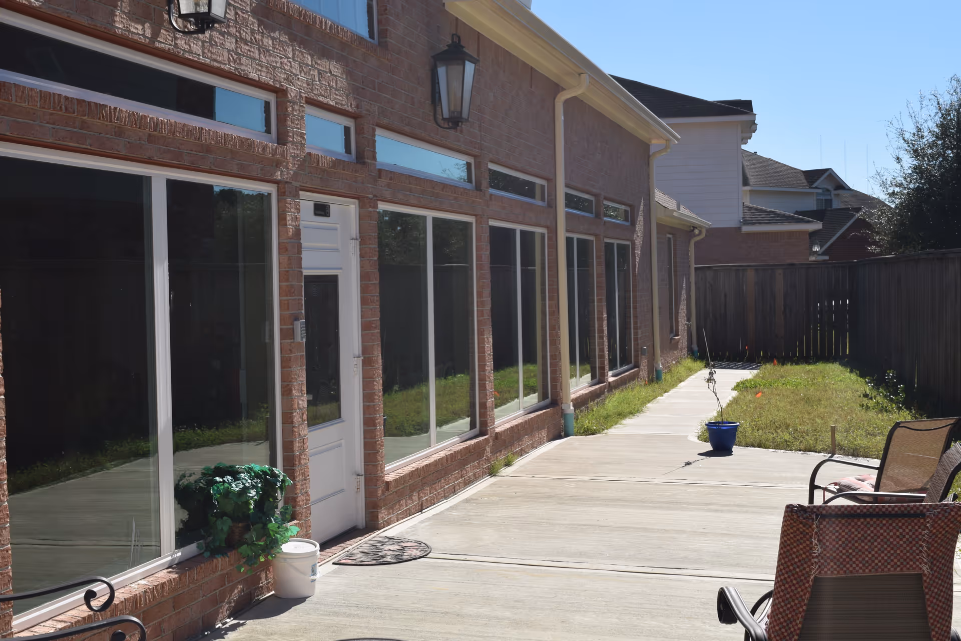 Outdoor patio area alongside a brick building with large windows and a white door. The patio has a concrete walkway, some potted plants, and outdoor chairs. A wooden fence and some grass are visible in the background under a clear blue sky.