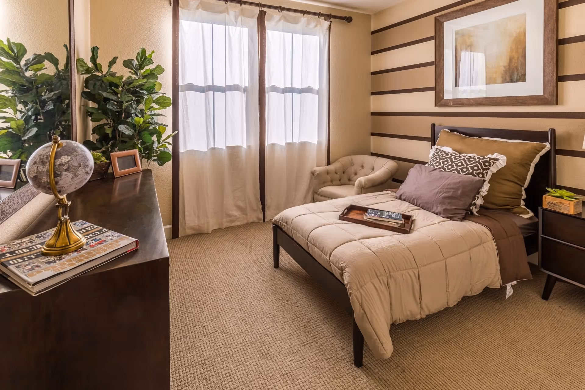 A cozy bedroom with a single bed covered in beige and brown bedding, a small tufted armchair in the corner, a dark wooden dresser with a globe and picture frames, and a large window with sheer curtains letting in natural light. The walls have a striped pattern and a framed artwork hangs above the bed.
