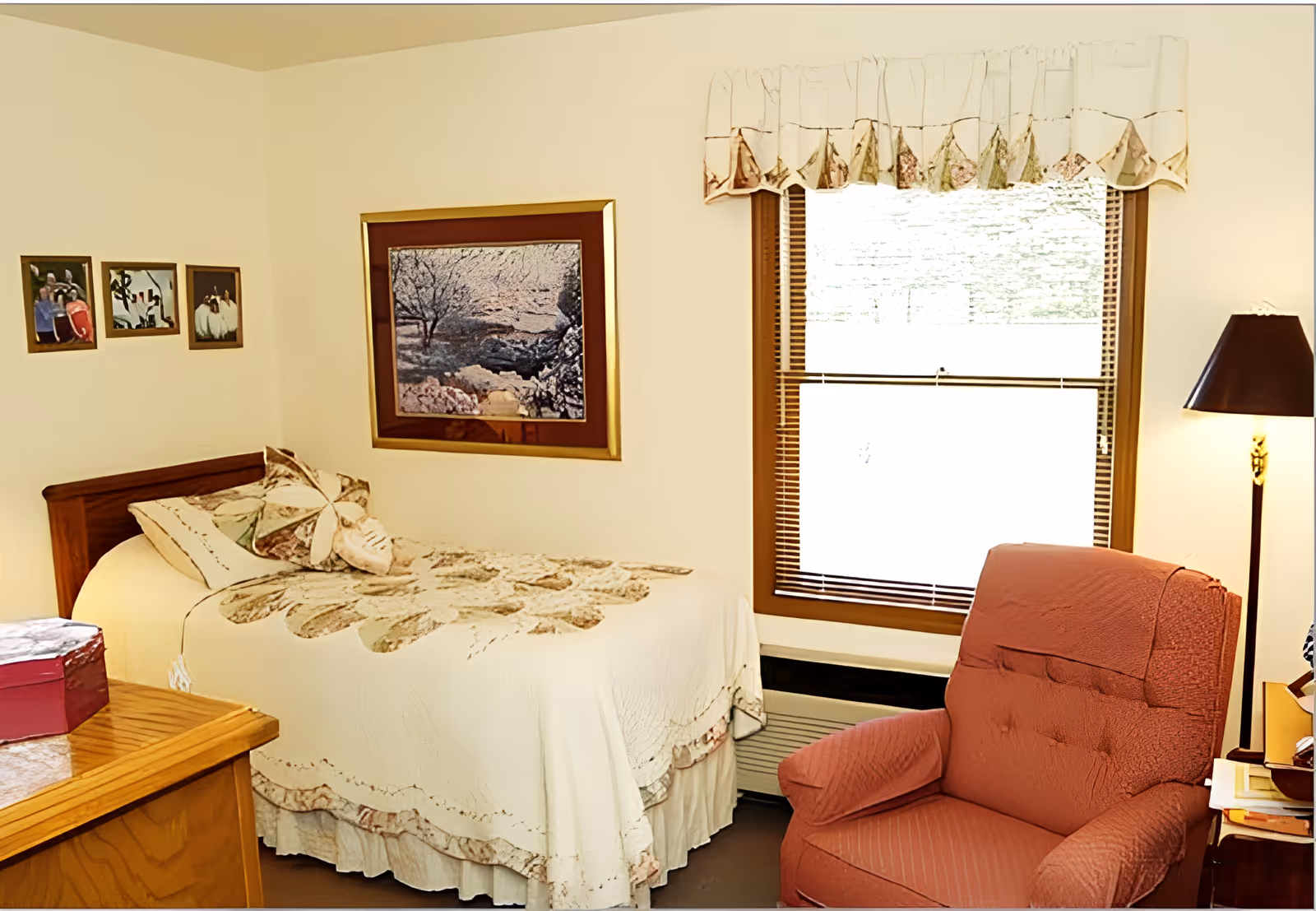 A cozy bedroom with a single bed covered in a cream-colored quilt with leaf patterns. There is a wooden headboard and a matching wooden dresser with a red box on it. A large window with wooden blinds and a decorative valance lets in natural light. Next to the window is a comfortable red armchair and a floor lamp. The walls are decorated with framed pictures and a large painting of a nature scene.