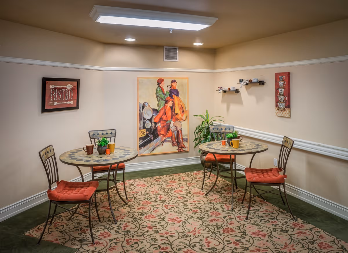 A small dining area with two round mosaic tables, each surrounded by two metal chairs with red cushions. The room has beige walls decorated with framed artwork and small shelves holding teacups. A patterned rug covers the green carpeted floor, and a potted plant is placed in the corner under soft ceiling lighting.