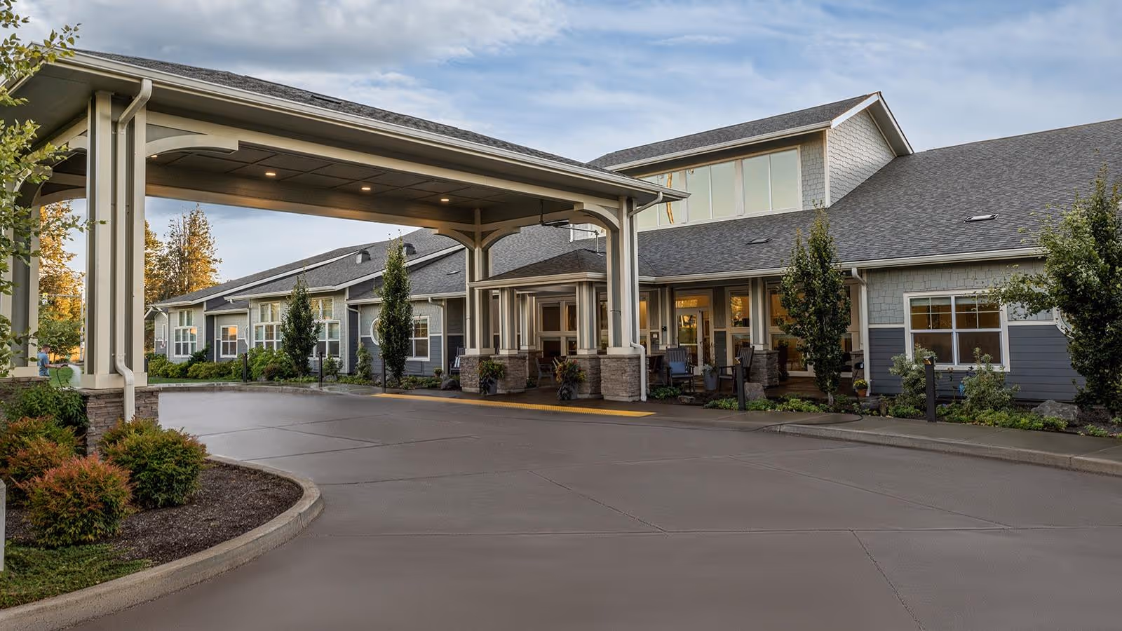 Exterior view of a senior living facility named Battle Creek Memory Care, showing a covered entrance with a driveway, landscaped bushes, and a large building with multiple windows under a cloudy sky.