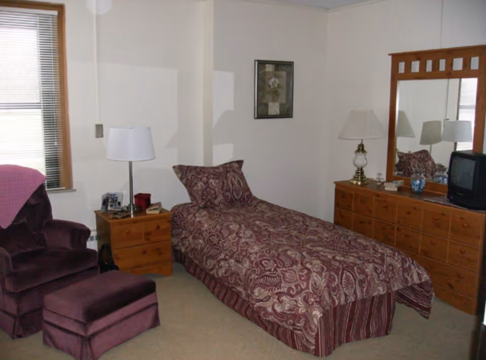 Small furnished bedroom with a single bed covered in a patterned burgundy bedspread, a wooden dresser with mirror and TV, a nightstand with lamp, and a purple armchair with ottoman.