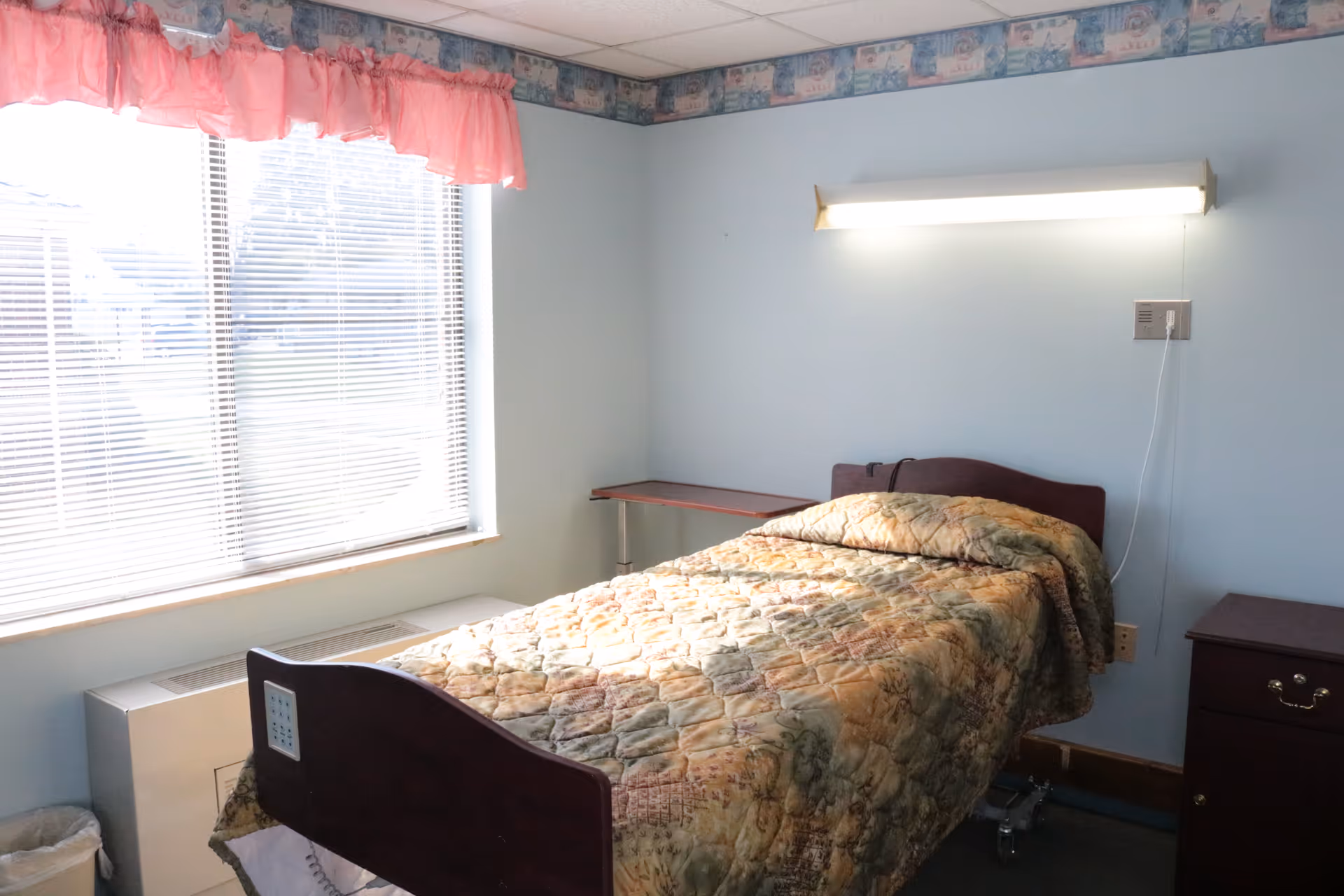 A small bedroom with a single bed covered in a patterned quilt. The room has light blue walls, a window with blinds and a pink valance, a wall-mounted light above the bed, a small wooden nightstand, and a small table in the corner.