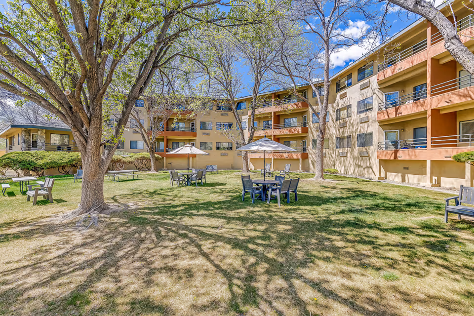 Outdoor courtyard area of a senior living facility with green grass, several trees, and multiple seating arrangements including tables with umbrellas and chairs. The building surrounding the courtyard is three stories high with balconies and windows. The sky is blue with some clouds.