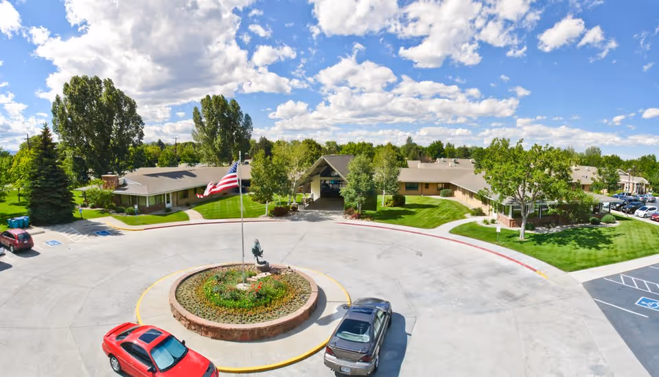Aerial view of North Shore Health and Rehabilitation Center showing a circular driveway with a landscaped roundabout featuring an American flag and a sculpture. The building is surrounded by green lawns, trees, and a parking area under a partly cloudy blue sky.