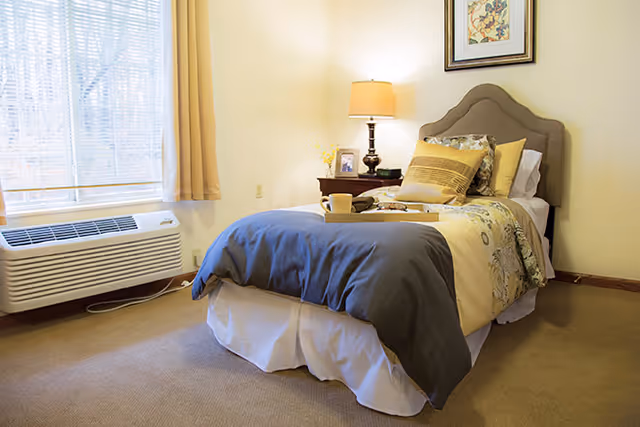 A cozy bedroom with a single bed featuring a gray and yellow comforter, multiple pillows, and a tray with a cup on it. There is a nightstand with a lamp, a framed photo, and a decorative item. A window with sheer curtains and yellow drapes is on the left side, and a wall-mounted air conditioning unit is below the window.