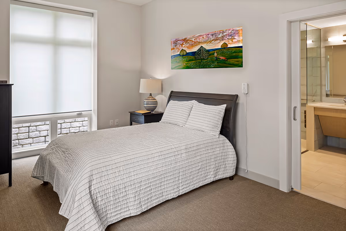 A neatly made bed with white and gray striped bedding in a bedroom. There is a nightstand with a lamp beside the bed, a colorful landscape painting on the wall above the headboard, and a large window with a white shade. An open door reveals a bathroom with a sink and mirror.