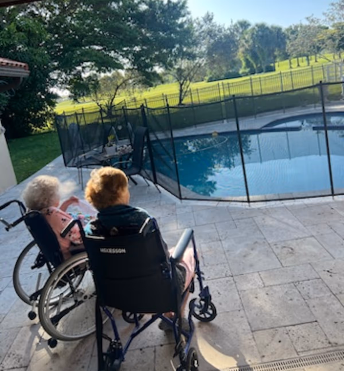 Two residents in wheelchairs sit on a tiled patio beside a fenced swimming pool with a grassy lawn and trees beyond.