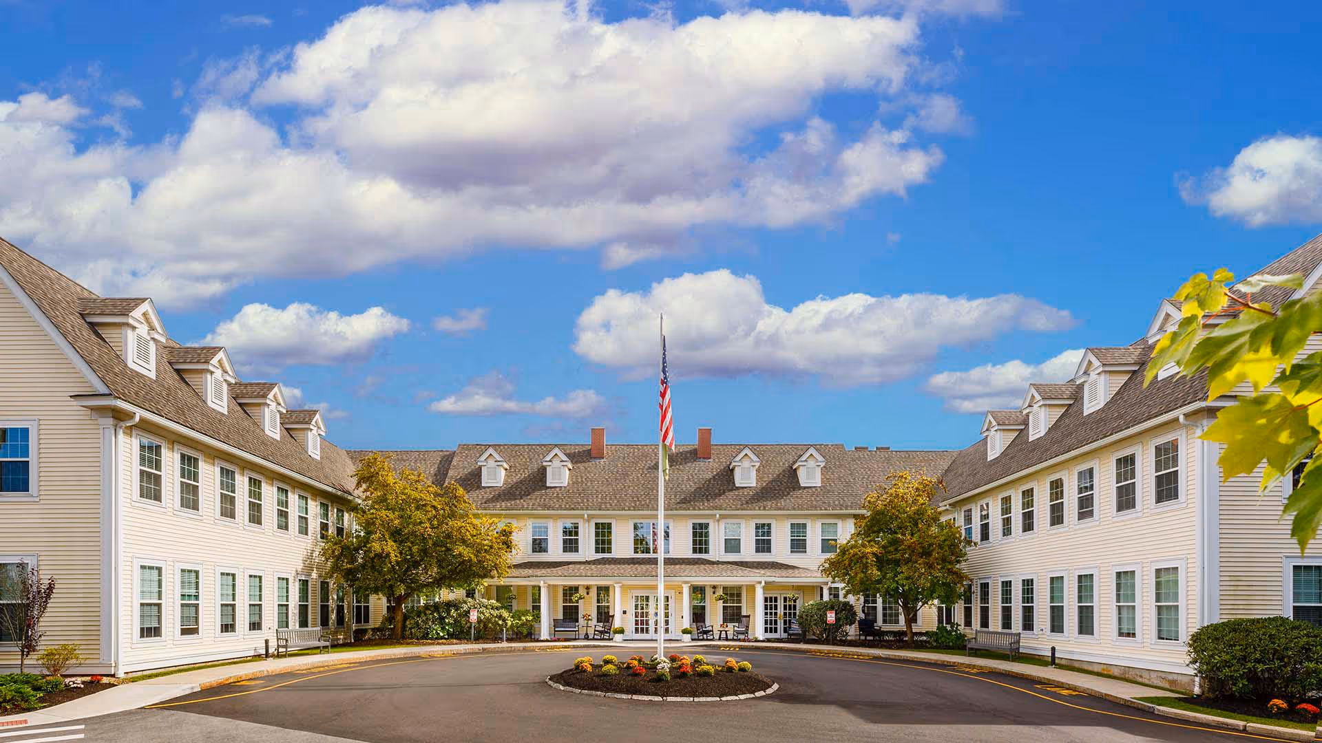 Front exterior view of Atria Merrimack Place, a large two-story building with white siding and multiple windows. The building forms a U-shape around a circular driveway with a flagpole and landscaped flower bed in the center. Trees and shrubs are planted near the building, and the sky is partly cloudy with blue patches.