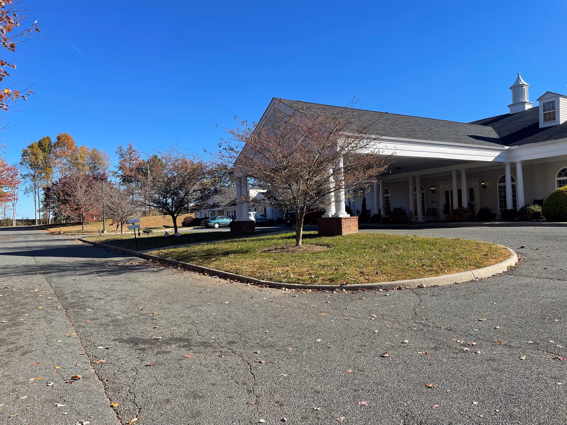 Exterior view of a senior living facility building with a covered entrance supported by white columns. There are small trees with autumn leaves and a clear blue sky in the background. The driveway and parking area are visible in the foreground.