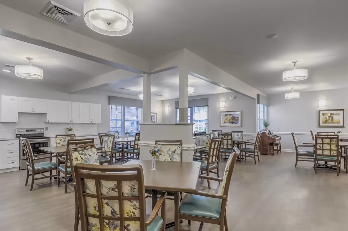 A bright and spacious dining room with multiple wooden tables and chairs featuring floral upholstery. The room has light-colored walls, several windows letting in natural light, modern ceiling lights, and a small kitchen area with white cabinets and a stove in the background. There is also a piano against one wall and framed artwork hanging on the walls.
