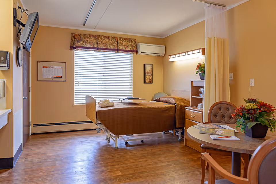 A well-lit patient room with a hospital bed covered in a brown blanket, a bedside table with a lamp and flowers, a round table with chairs and a flower arrangement, a window with blinds and a decorative valance, and a wall-mounted television.