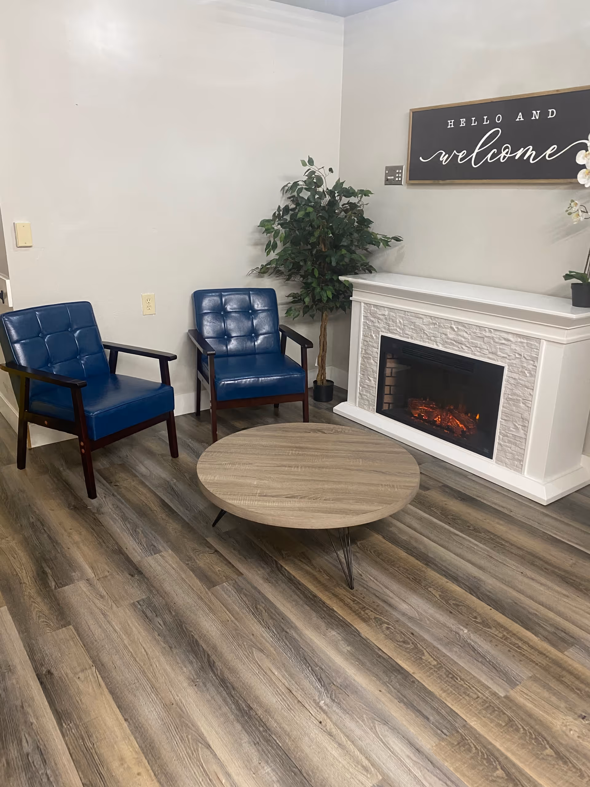 A cozy sitting area with two blue cushioned chairs, a round wooden coffee table, a white electric fireplace, a potted plant, and a wall sign that reads 'Hello and welcome'. The floor is covered with wood-patterned vinyl planks.