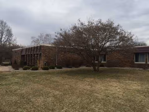 Brick-assisted living facility facade with a lawn, shrubs, and a central tree.