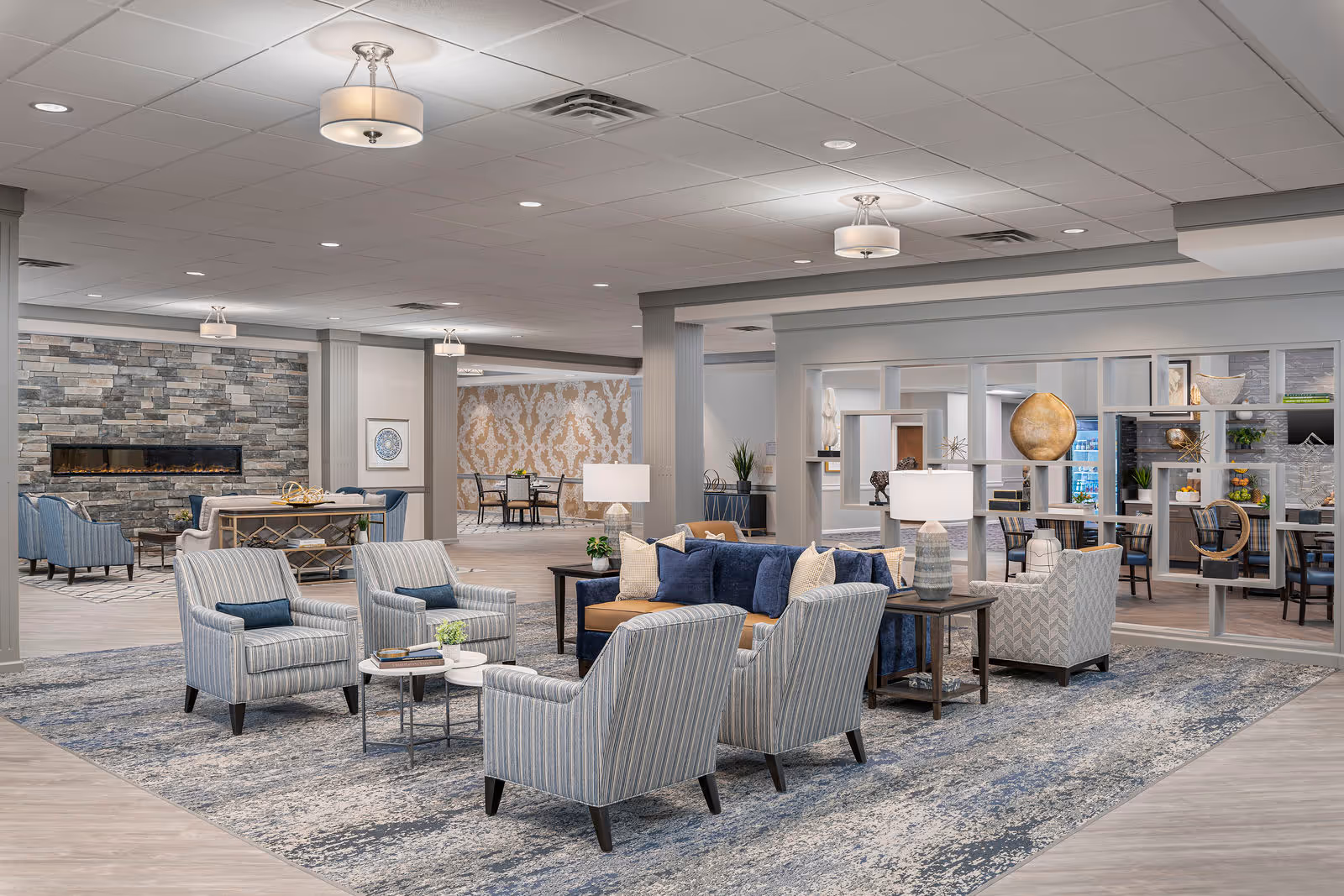 A spacious and elegantly furnished senior living facility common area with multiple seating arrangements including striped armchairs and a blue sofa around coffee tables on a large patterned rug. The room features modern ceiling lights, a stone fireplace, decorative shelving with art pieces, and a dining area visible in the background with patterned wallpaper.