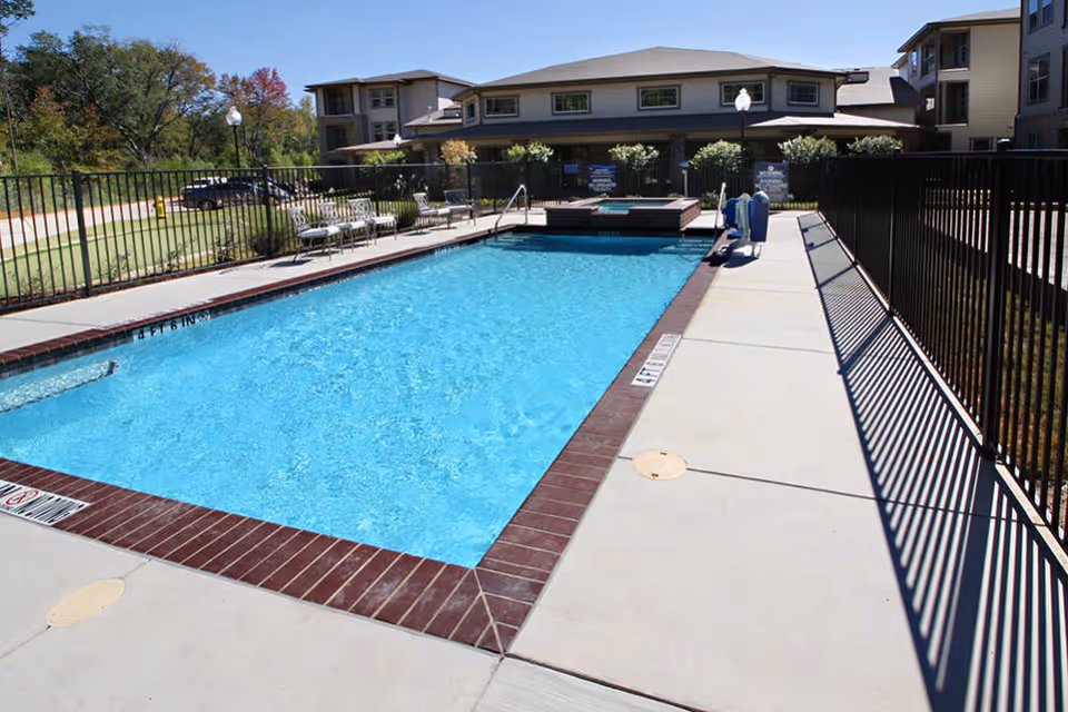 A fenced outdoor swimming pool with lounge chairs and a hot tub beside a multi-story senior living building.