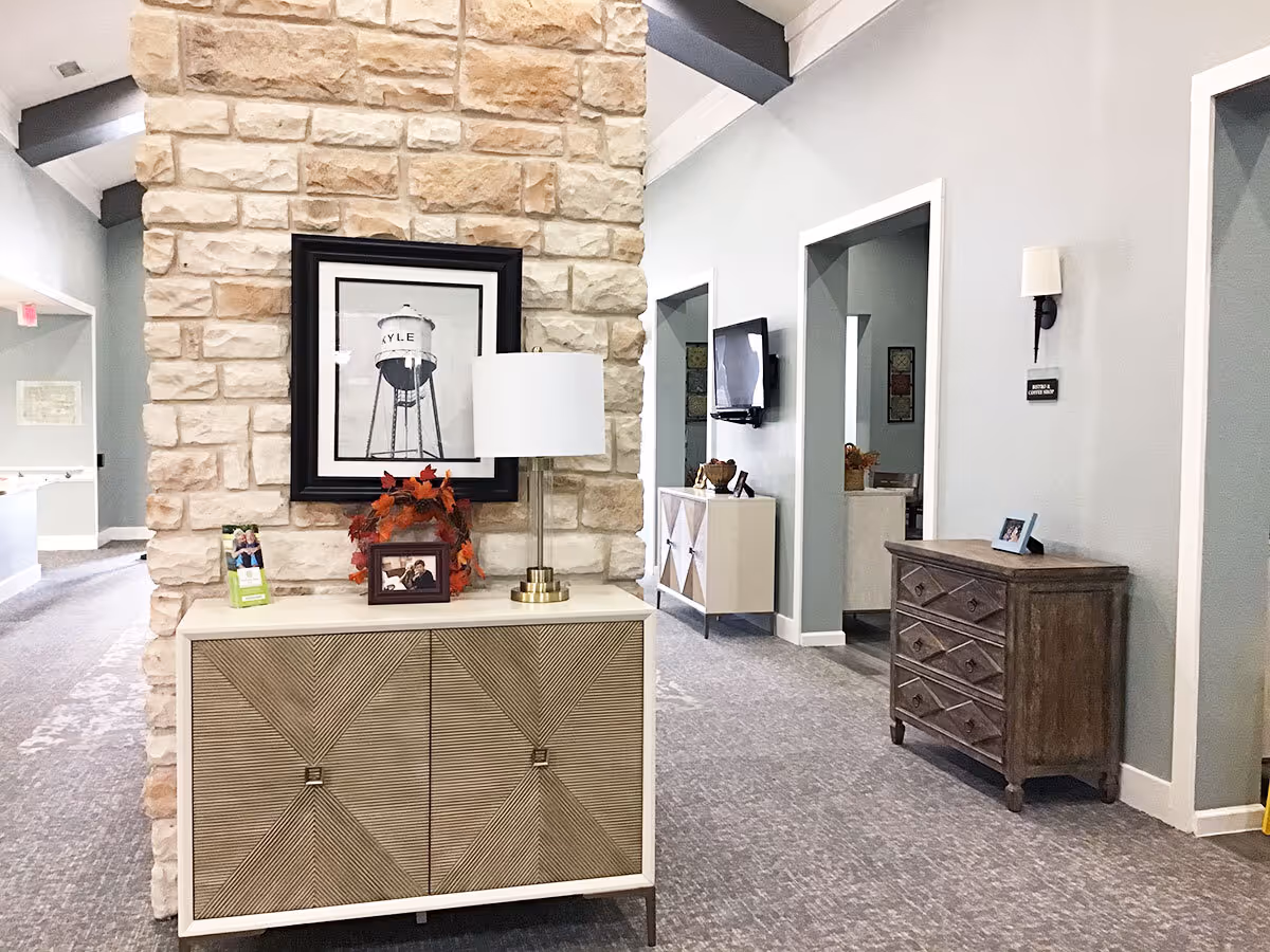 Interior hallway of Orchard Park of Kyle Assisted & Senior Living featuring a stone accent wall with a framed picture of a water tower labeled 'KYLE'. In front of the wall is a decorative cabinet with a table lamp, a small framed photo, and a fall-themed wreath. The hallway has light blue walls, carpeted floors, and additional furniture including a wooden chest of drawers and a wall-mounted TV.