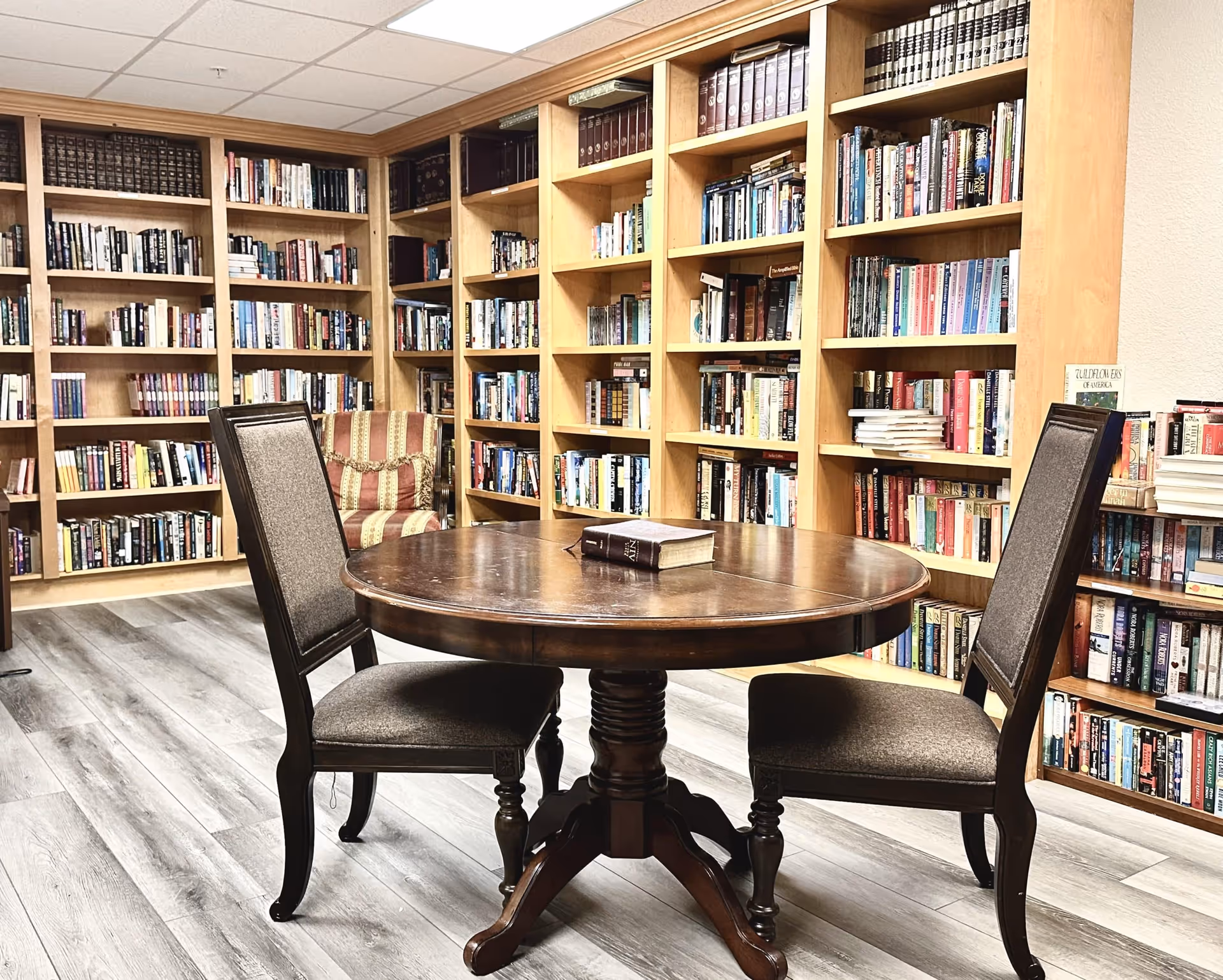A cozy library room with wooden bookshelves filled with books lining the walls. In the center, there is a round wooden table with two upholstered chairs on either side. A single book lies on the table. The floor has light-colored wood planks, and there is a striped armchair in the corner.