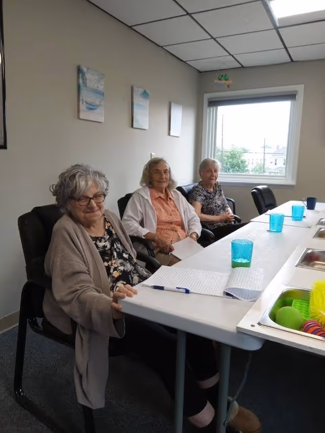 Three elderly women sitting in chairs around a long table in a well-lit room with a window. The table has papers, pens, and blue drinking glasses on it. The room has light-colored walls with three small paintings and a ceiling with fluorescent lights.