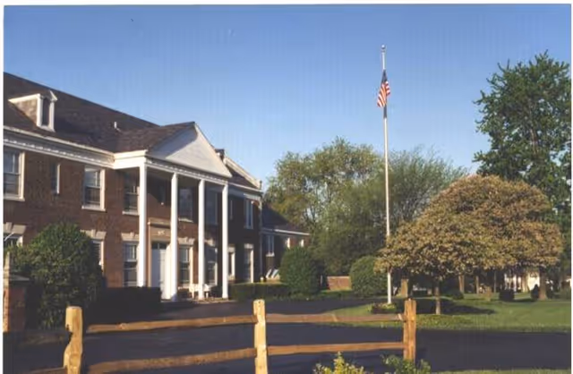 Exterior view of a two-story brick building with white columns at the entrance, surrounded by trees and bushes. An American flag is flying on a flagpole in front of the building, and a wooden fence is visible in the foreground under a clear blue sky.