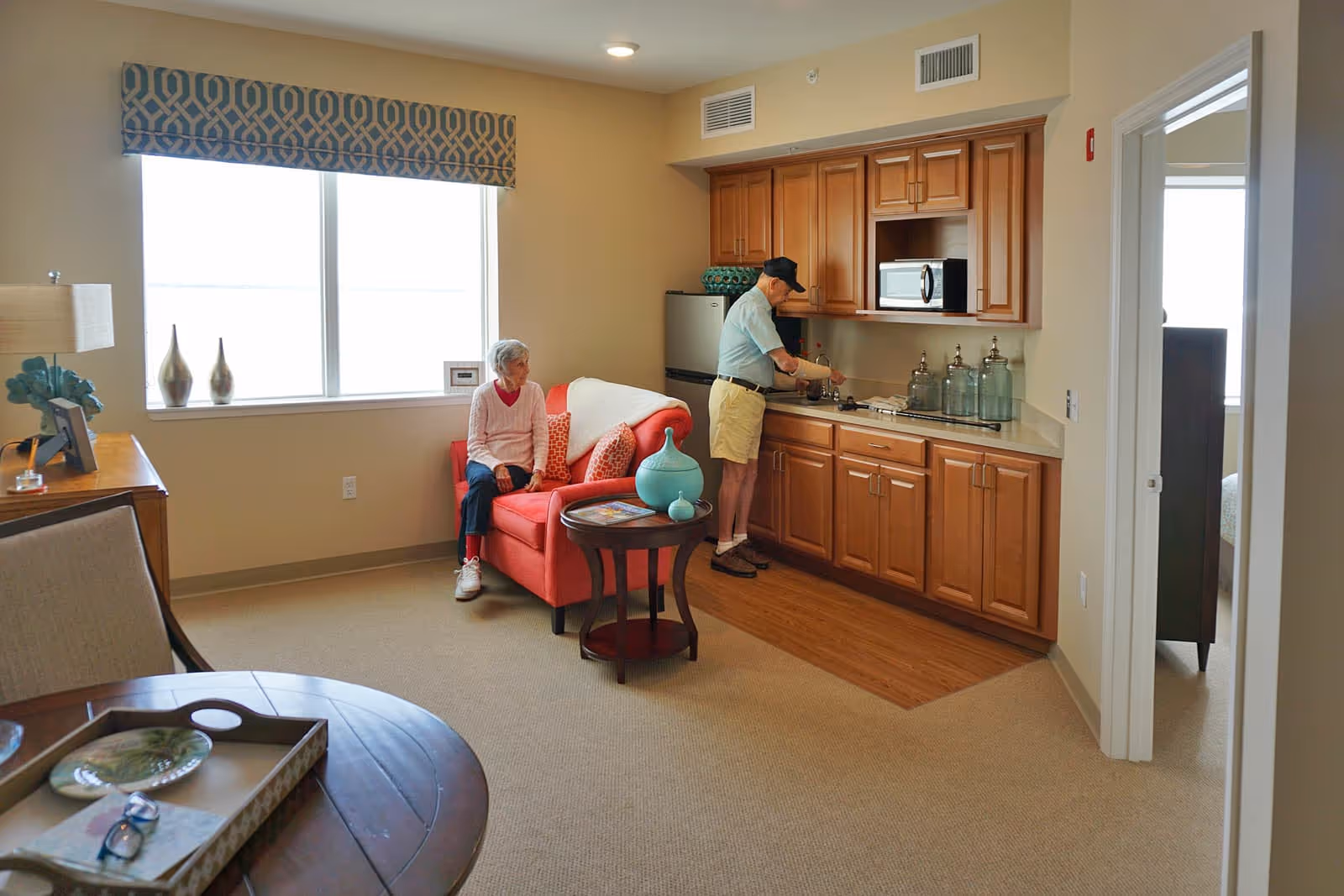 A senior man is preparing something at a kitchenette with wooden cabinets, a microwave, and a small refrigerator. A senior woman is sitting on a red loveseat with patterned cushions near a window with a decorative valance. The room has beige walls and carpet, with a round wooden table and a lamp on a side table.